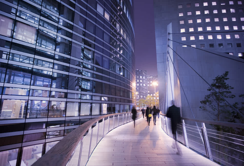 People walk along a curved, elevated walkway at night, surrounded by modern glass office buildings with illuminated windows. The scene is urban and futuristic, with blurred motion giving a sense of activity and movement.