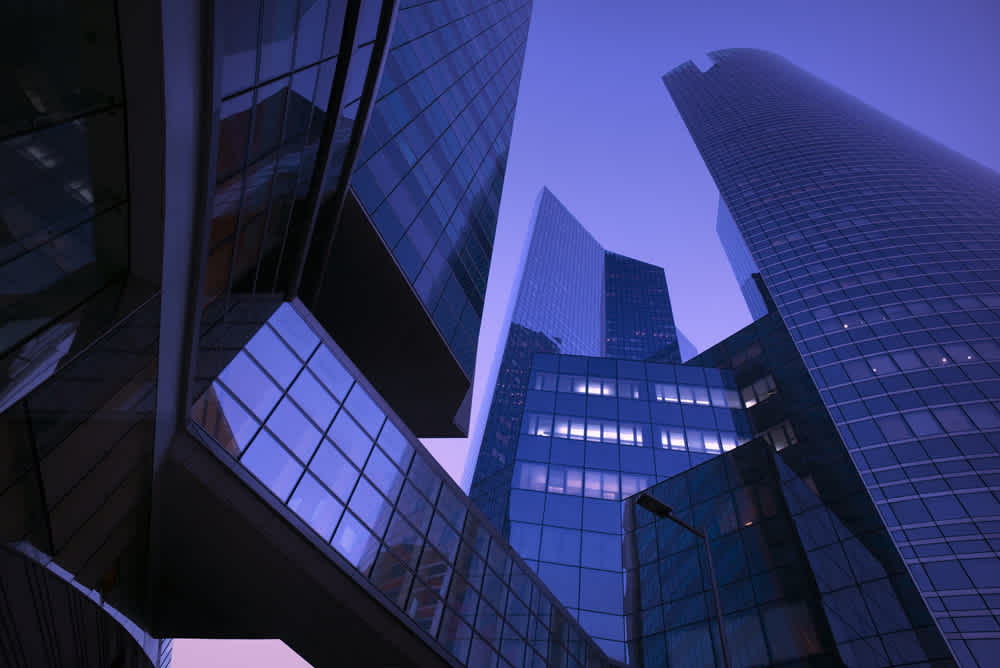 Low-angle view of modern glass skyscrapers under a dusk sky, reflecting shades of blue and purple. The buildings feature sleek, angular designs with interconnected walkways, emphasizing a futuristic architectural style.
