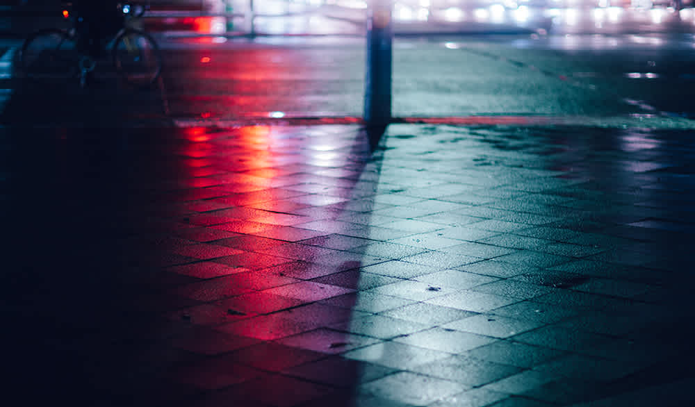Wet street tiles reflecting red and blue lights at night. A bicycle is partially visible on the left, and bright, blurred lights illuminate the background, creating an atmospheric urban scene.