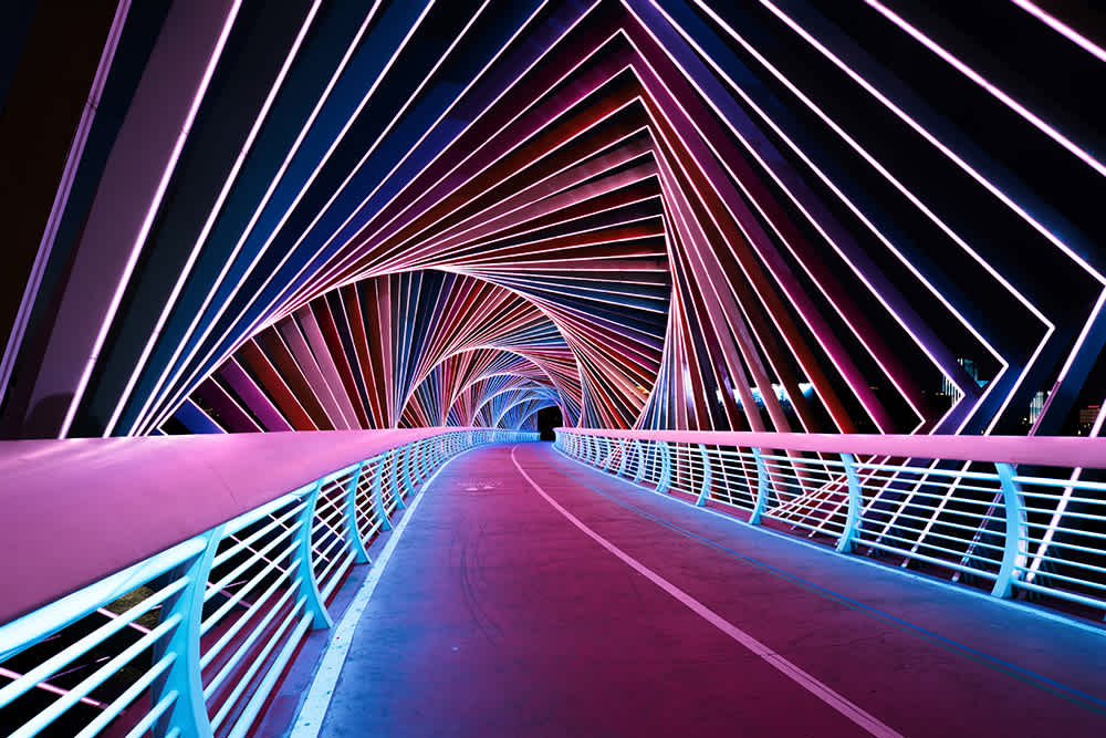 A futuristic, illuminated tunnel with vibrant, multicolored lights forming geometric patterns overhead. The curved pathway, flanked by railings, leads into the distance, creating a visually striking and dynamic scene.