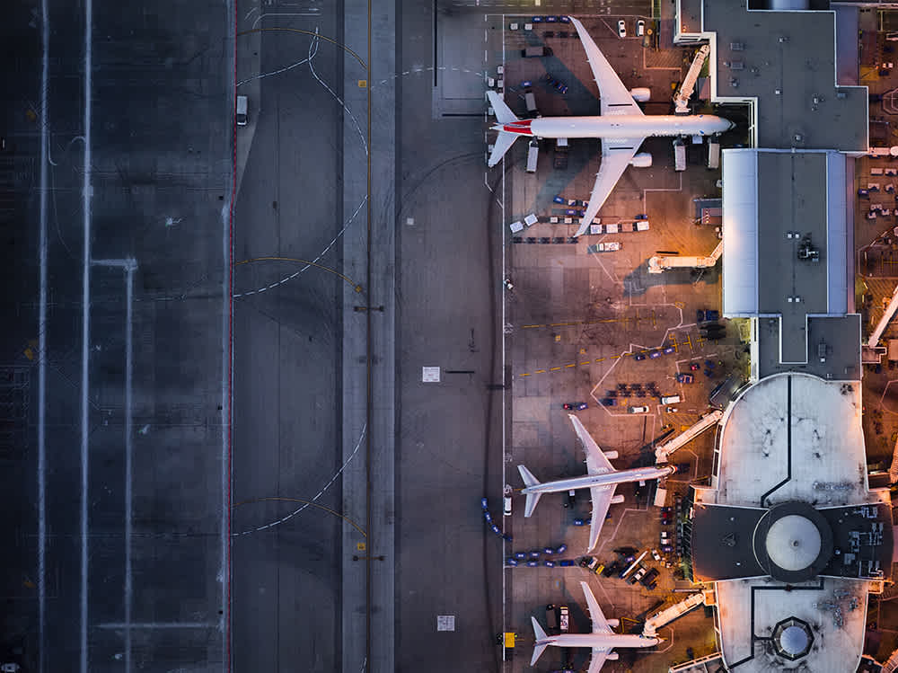Aerial view of an airport terminal at dusk: three airplanes are parked along the gates with vehicles around them, preparing for takeoff or unloading. The surrounding tarmac is illuminated by lights, casting long shadows.