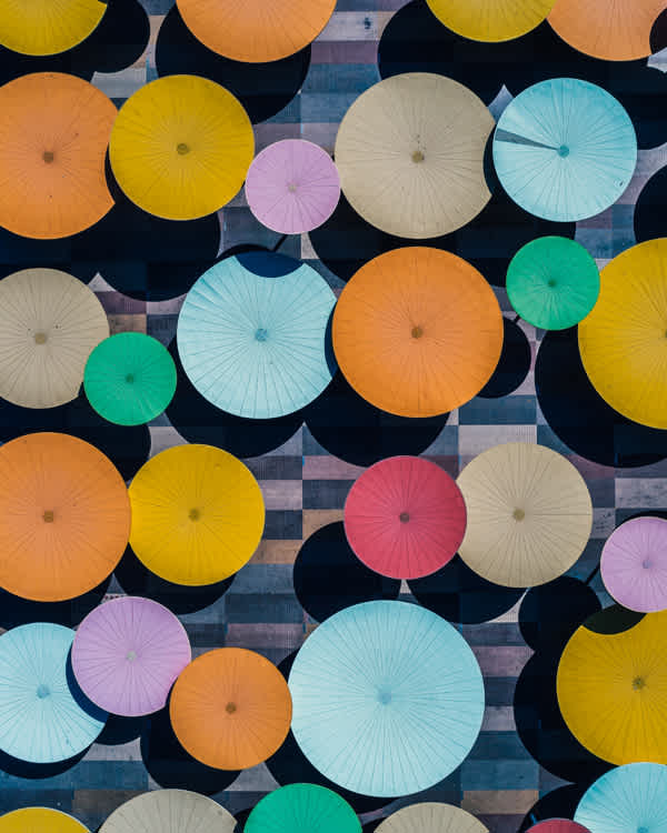 Aerial view of colorful umbrellas in shades of yellow, orange, pink, blue, and green, creating a vibrant pattern on a dark background with geometric shapes.