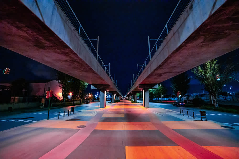 A vibrant, illuminated street scene at night, with a raised concrete structure running above. The ground below is painted in colorful blocks of orange, pink, and yellow. Streetlights and trees line the sides, creating a symmetrical view.