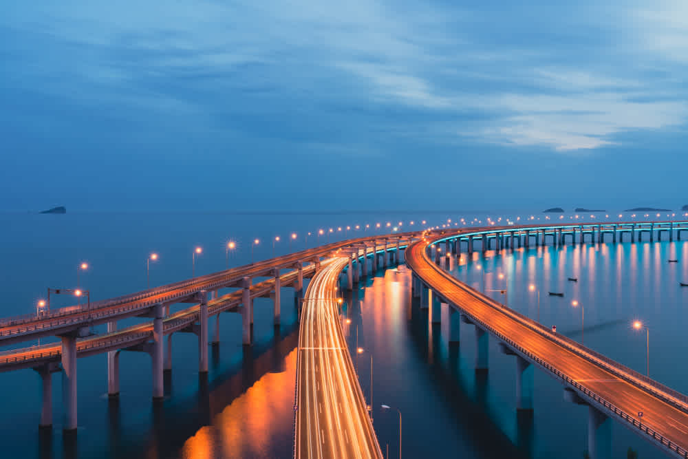 A long, curving bridge with glowing streetlights spans across calm water under a blue, cloudy sky during dusk. Light trails from vehicles create bright lines along the bridge, enhancing the serene and modern scene.