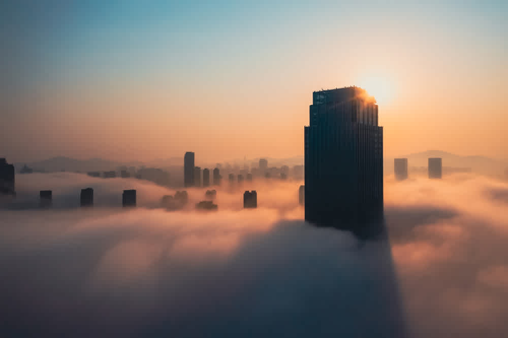 A city skyline at sunrise with sunlight illuminating a tall building. Dense fog envelops the lower parts of the buildings, creating a surreal atmosphere. The horizon shows distant hills under a colorful sky transitioning from blue to orange.