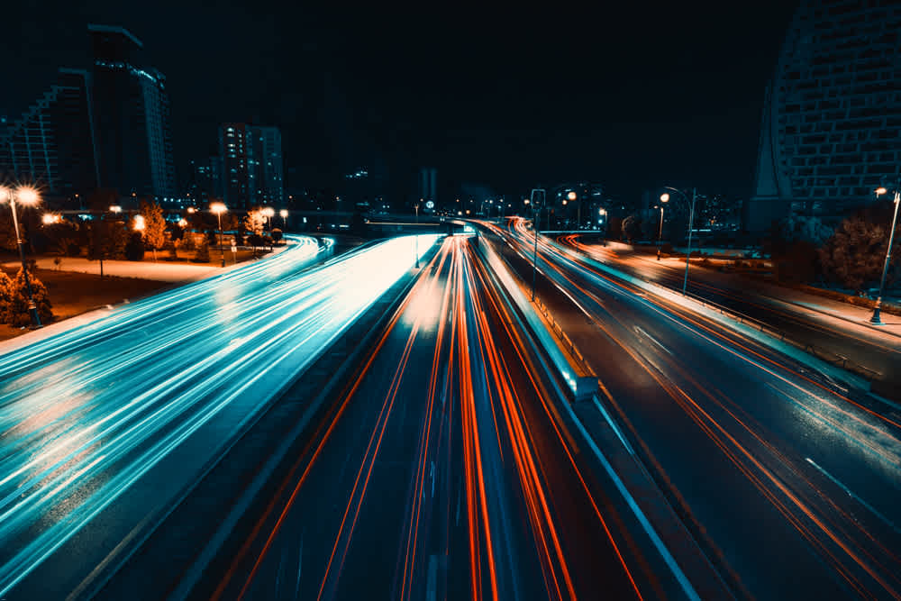 A nighttime long-exposure photo of a highway with light trails from vehicles. Blue and red streaks from car lights create dynamic lines against a dark urban backdrop, with buildings and streetlights visible in the distance.