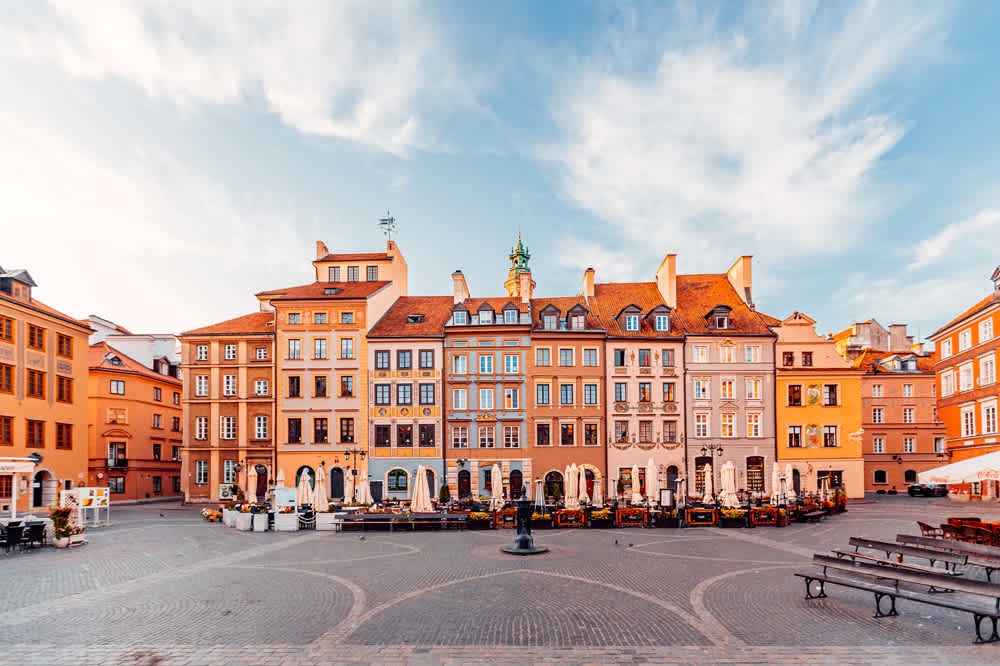 A wide-angle view of a historic European town square featuring colorful, ornate buildings with gabled roofs and a central clock tower. The square has outdoor cafes and cobblestone paving, with benches and a clear blue sky above.