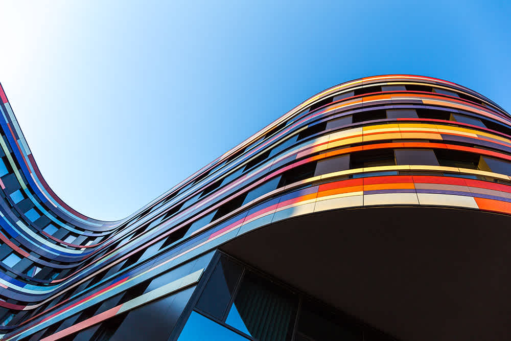 Curved modern building with colorful horizontal stripes in red, orange, yellow, and blue, stands against a clear blue sky. The structure features smooth, wavy lines and reflective glass elements.