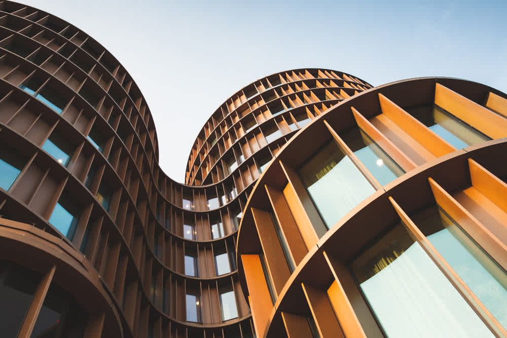 Curved, modern building with a unique facade featuring vertical, rust-colored metal panels and large, reflective windows. The structure rises against a clear blue sky, showcasing its contemporary architectural design.