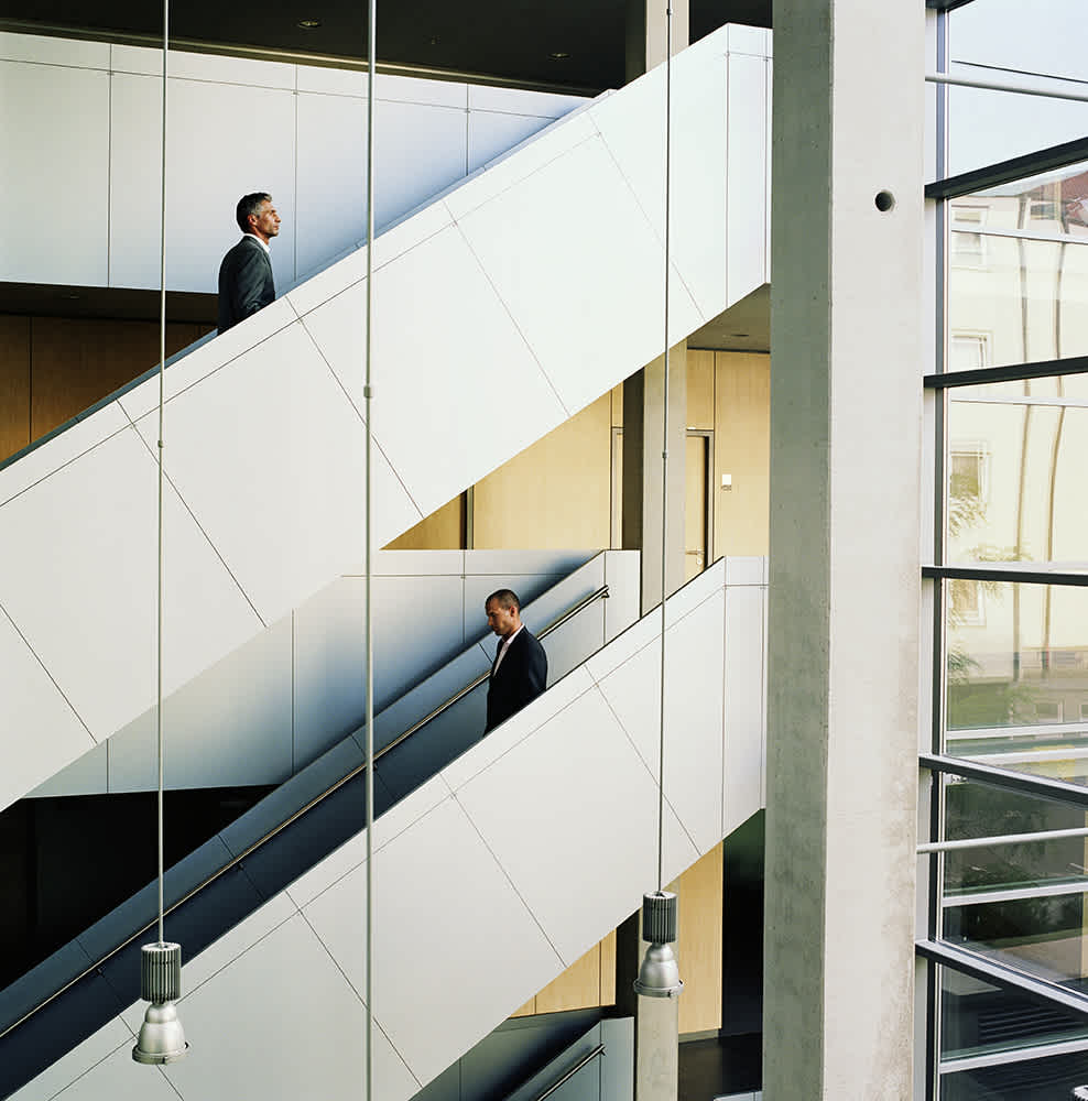 Two people in suits walk on parallel escalators in opposite directions inside a modern building. Large windows allow natural light to illuminate the space. Hanging lights and a concrete pillar add to the contemporary design.