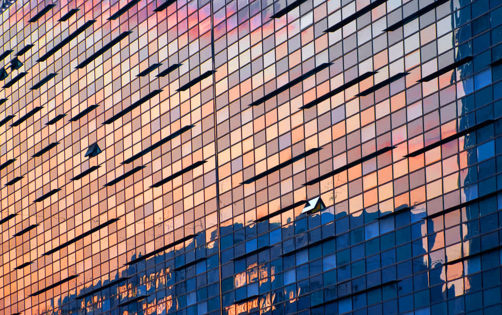 A glass skyscraper reflecting a colorful sunset. The buildings facade has irregular horizontal lines, creating a dynamic pattern. The sky and clouds create hues of pink, orange, and blue across the mirrored surface.
