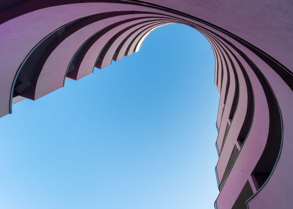 A view from the bottom of a modern building with pink curving balconies, looking up at a clear blue sky. The structure forms a wave-like pattern around the open sky, creating an abstract and symmetrical visual effect.