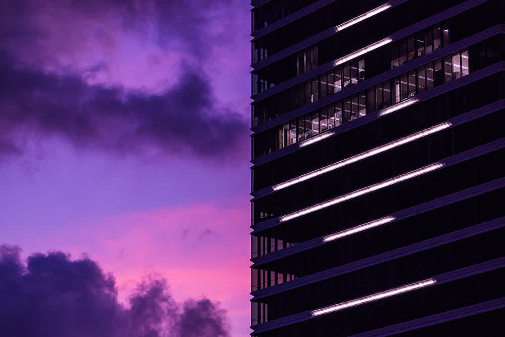 A modern building with illuminated windows stands against a dramatic sky at dusk. The sky features vibrant purple and pink hues, with dark clouds creating a striking contrast.
