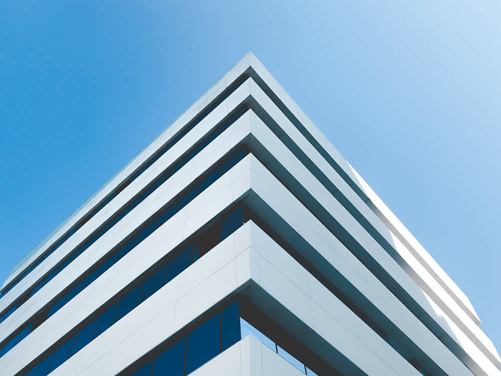 Corner view of a modern, angular building with reflective windows against a clear blue sky. The architecture features clean, white lines and geometric shapes, creating a minimalist and contemporary appearance.