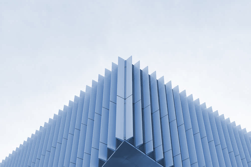 Photo of a modern building with a facade of vertical, evenly spaced blue-gray fins, creating a geometric pattern. The sky is overcast, providing a muted contrast to the clean lines and angular shapes of the architectural design.