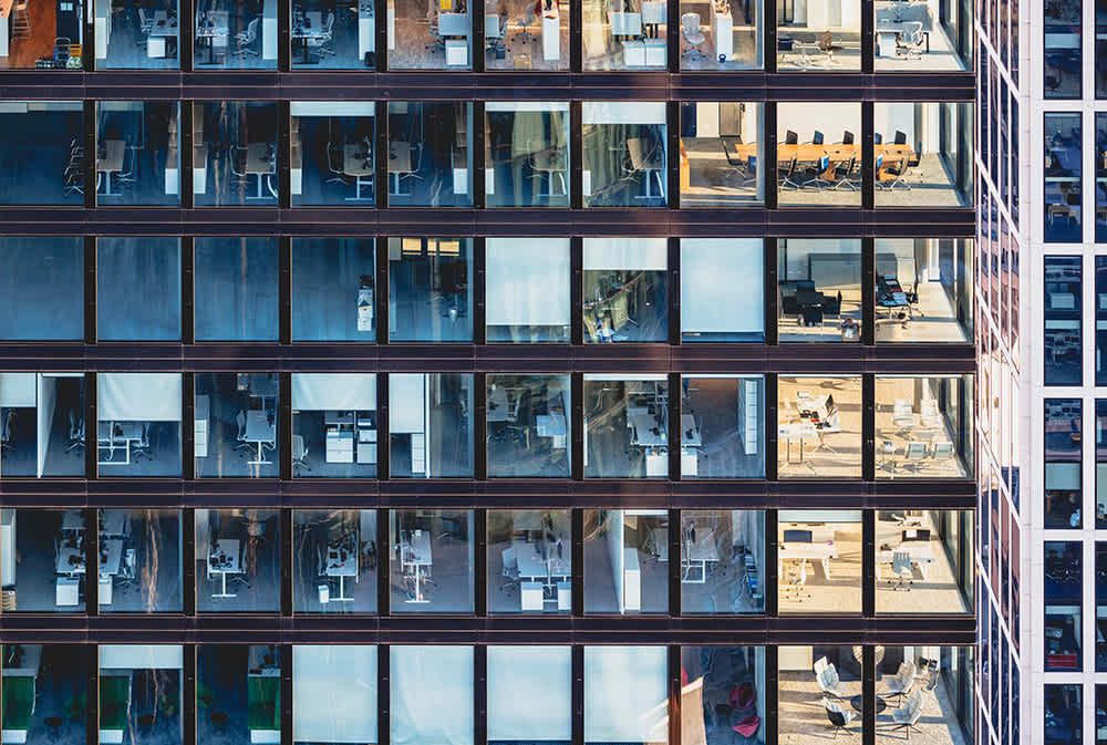 Exterior view of a high-rise office building with large glass windows revealing multiple floors. Inside, rows of desks, office chairs, meeting rooms, and a few people are visible, suggesting a typical modern workplace environment.