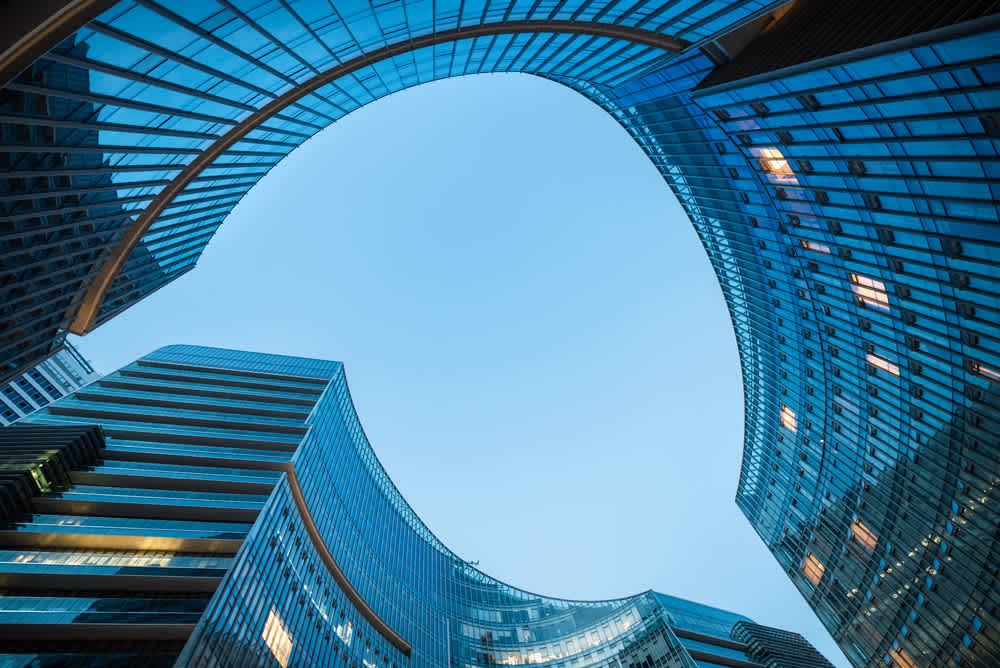 Curved modern glass buildings form an open oval shape against a clear blue sky. The facades reflect light, creating a dynamic and futuristic appearance. The view is from ground level, looking upwards.