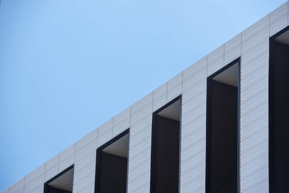 A modern building facade with vertical, rectangular windows and a light gray surface against a clear blue sky. The windows create a pattern of shadows, giving depth and contrast to the geometric design.