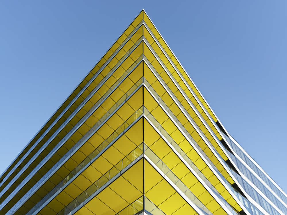 A modern building with bold yellow angular lines against a clear blue sky. The perspective highlights the sharp geometric design, emphasizing the symmetry and contrasts of the structure.