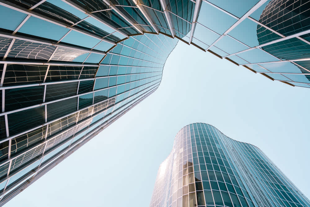 Upward view of modern skyscrapers with curved glass facades against a clear blue sky. The architecture reflects light, creating a sleek and dynamic composition.