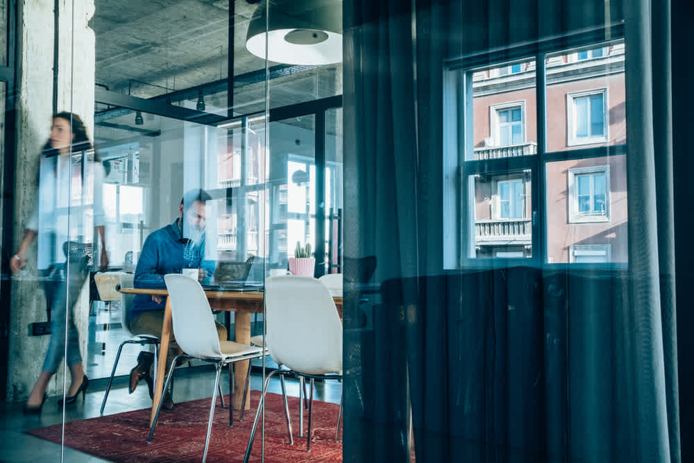 A modern office with large windows showing a red brick building outside. Inside, a person sits at a desk with laptops, while another walks past. The room has a mix of industrial decor, white chairs, and a red rug.
