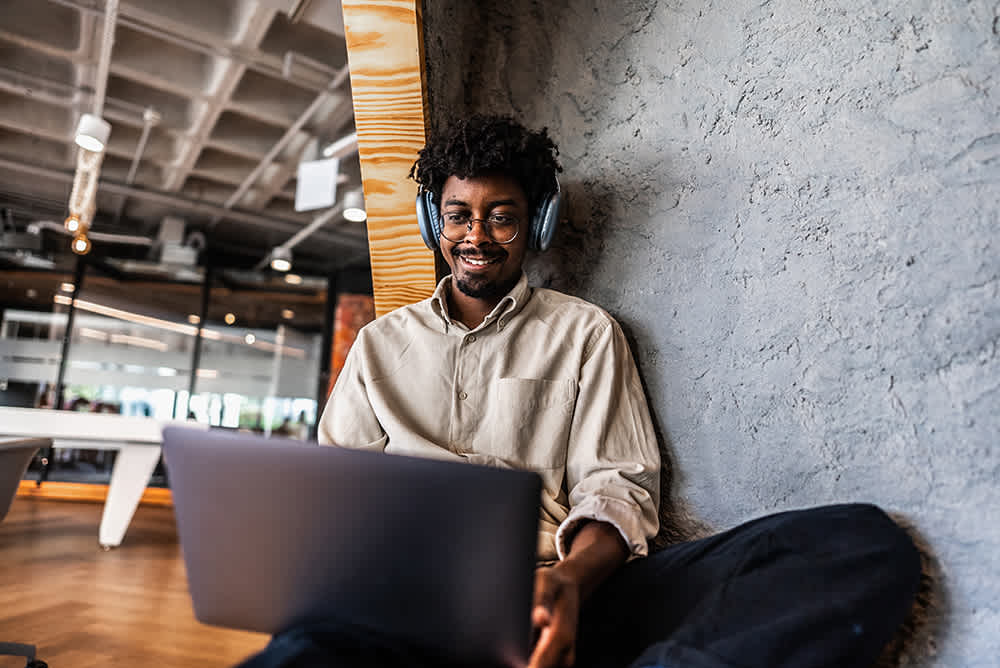 A person is sitting on the floor against a stone wall, wearing headphones and smiling while using a laptop. The background features an open office space with large windows and modern furniture.