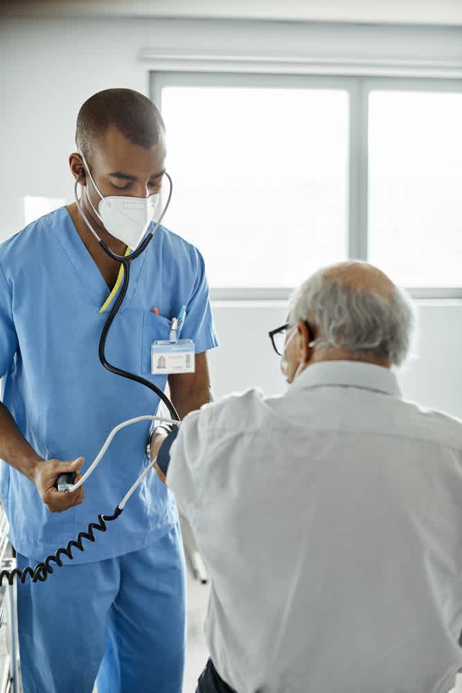 A healthcare worker in blue scrubs and a mask uses a stethoscope and blood pressure cuff to check an elderly mans blood pressure. The setting appears to be a medical facility with a window in the background.