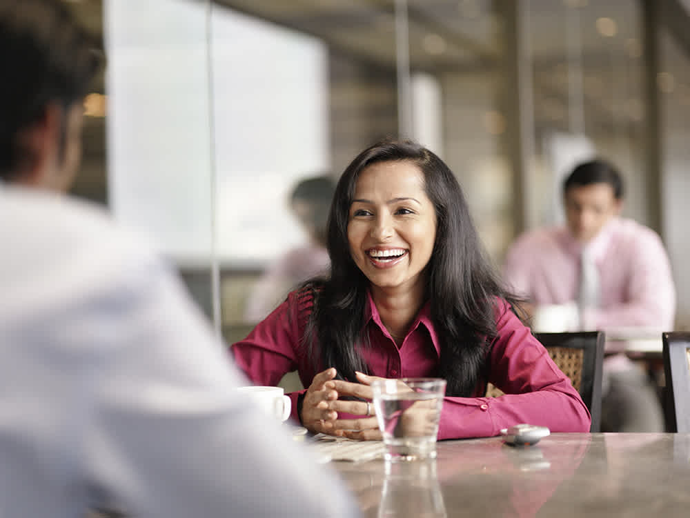 A woman in a pink shirt smiles while talking to a man across a table in a cafe. A glass of water and a cup are on the table. Two people in similar attire are blurred in the background.