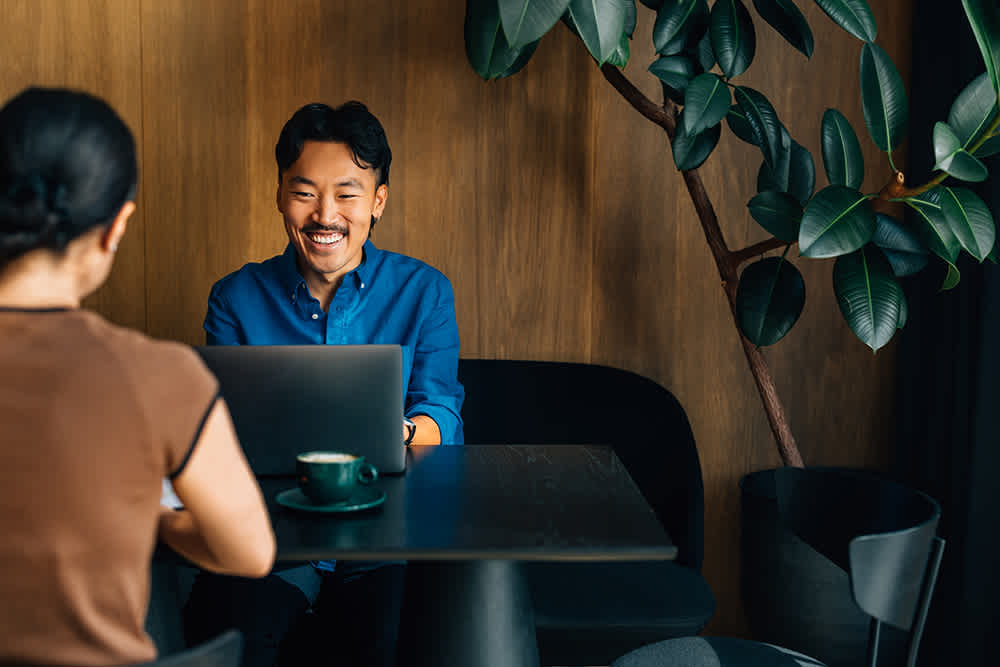 Asian man with mustache, in blue shirt, working at a coffee shop next to a plant