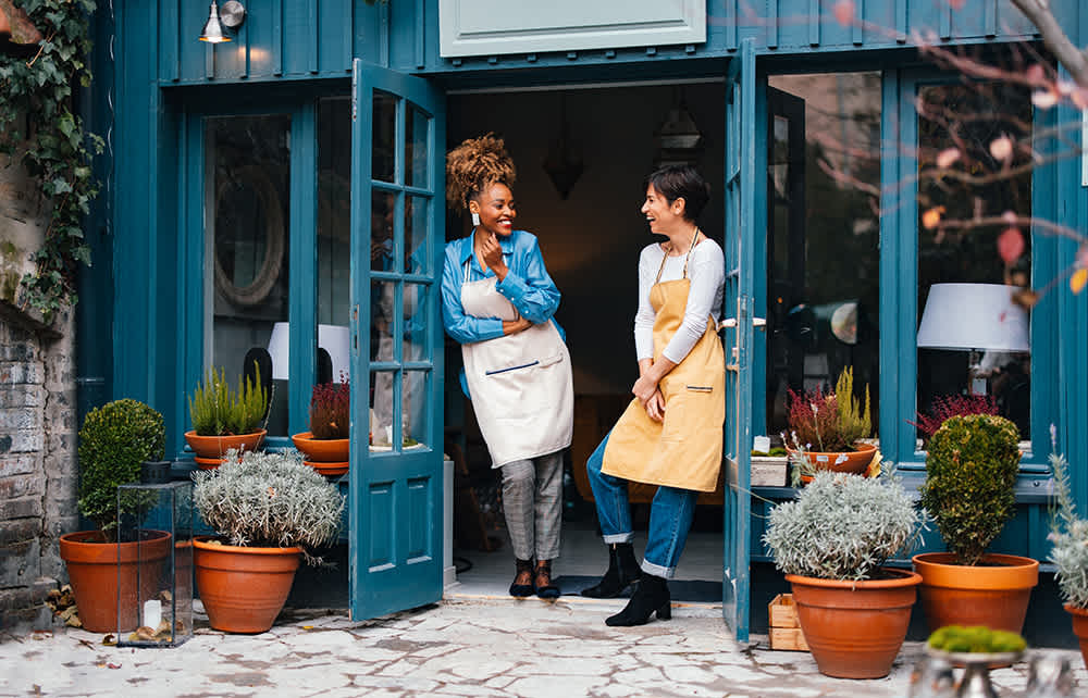 Two people are smiling and chatting at the open entrance of a cozy blue storefront. They both wear aprons, one blue and one yellow. Potted plants line the doorway, creating a welcoming atmosphere.