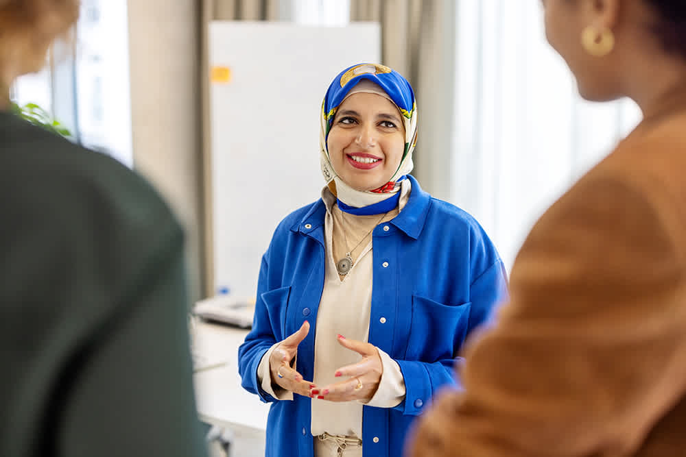 A smiling woman in a blue jacket and patterned headscarf gestures while talking to two people. She stands in a bright room with a whiteboard in the background.
