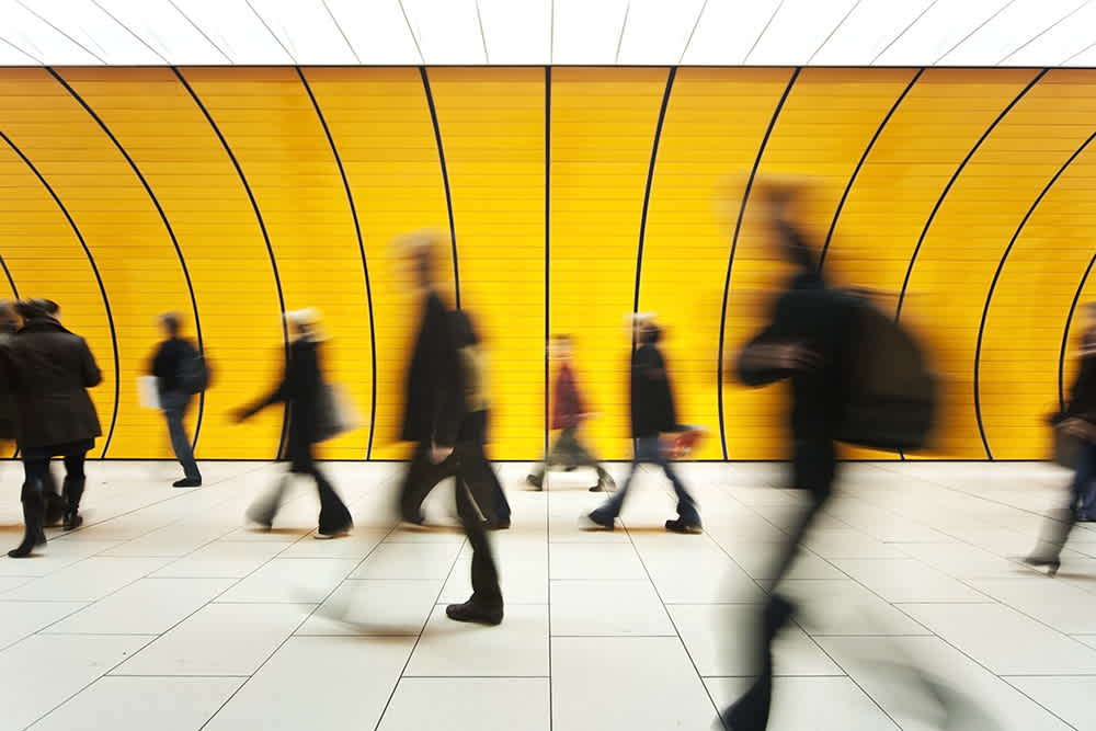 Blurred image of people walking in a hallway with a vibrant yellow wall and curved lines in the background. The floor is tiled, and the motion gives a sense of hustle and bustle.