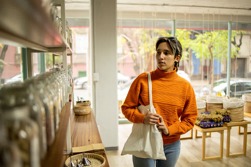 A person in an orange sweater shops in a bulk goods store, holding a cloth bag and looking at jars on a shelf. Sunlight streams through large windows, illuminating baskets and the store interior.