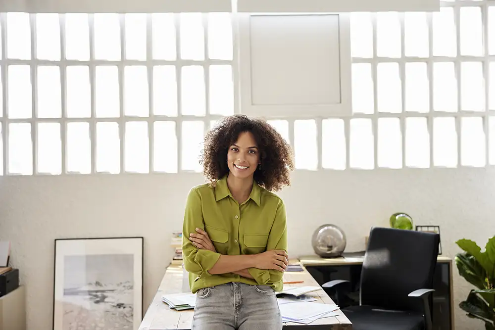 Black woman in green shirt and jeans leaning against her office desk