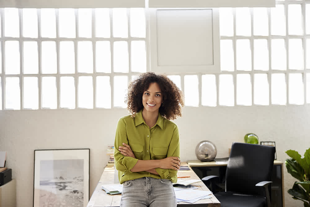 Black woman in green shirt and jeans leaning against her office desk