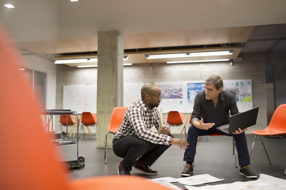 Two men are crouched on the floor of a modern office, discussing documents spread out in front of them. One holds a laptop, and empty orange chairs fill the background.