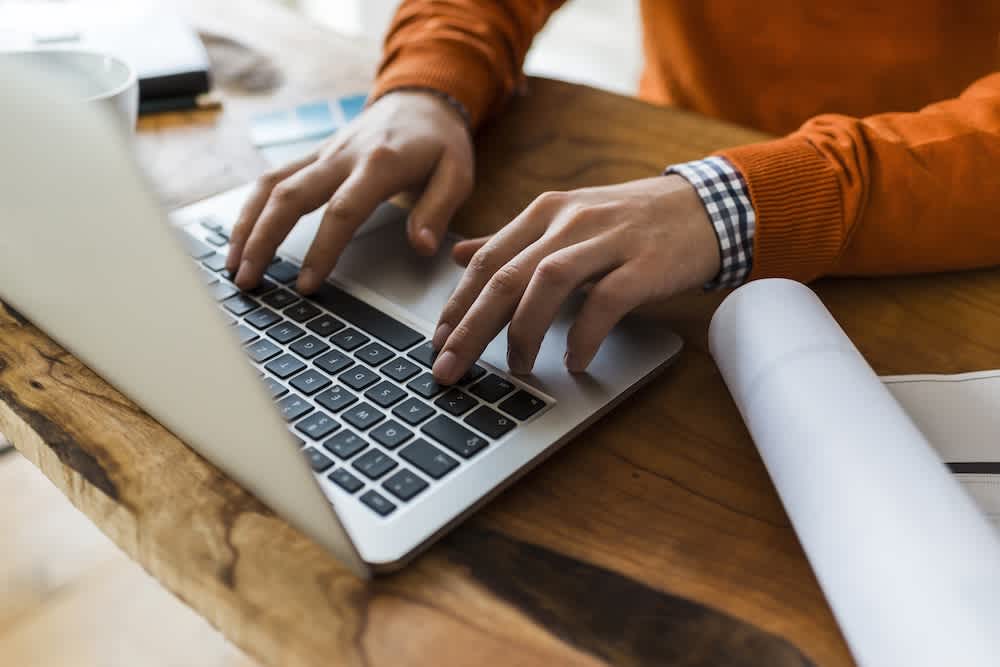 A person in an orange sweater types on a laptop at a wooden desk with a rolled-up paper and a smartphone nearby. Only their hands and torso are visible.