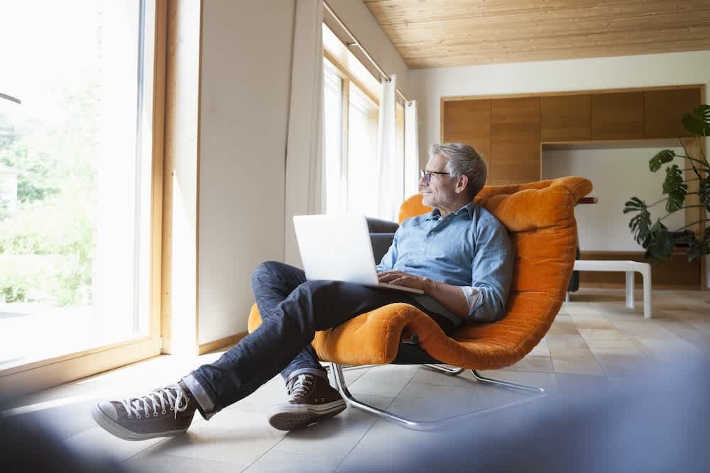 A man sits on an orange lounge chair with a laptop on his lap, looking out a large window in a modern, bright room with wooden accents and indoor plants.