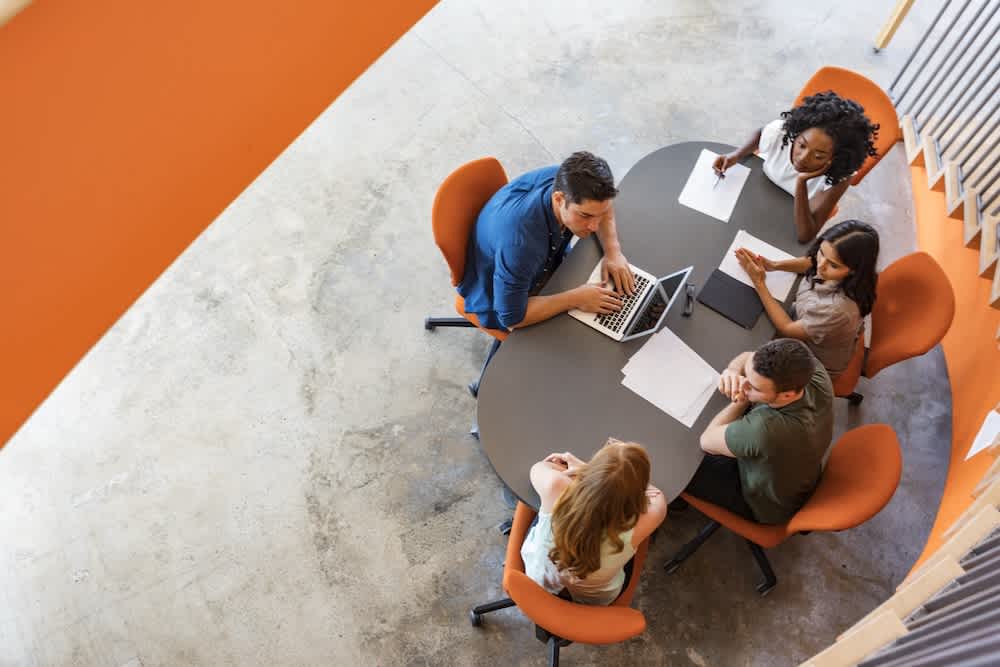 Five people sit around an oval table in an office, with papers and a laptop, engaged in discussion. The photo is taken from above, showing an informal meeting setting with orange chairs and a concrete floor.