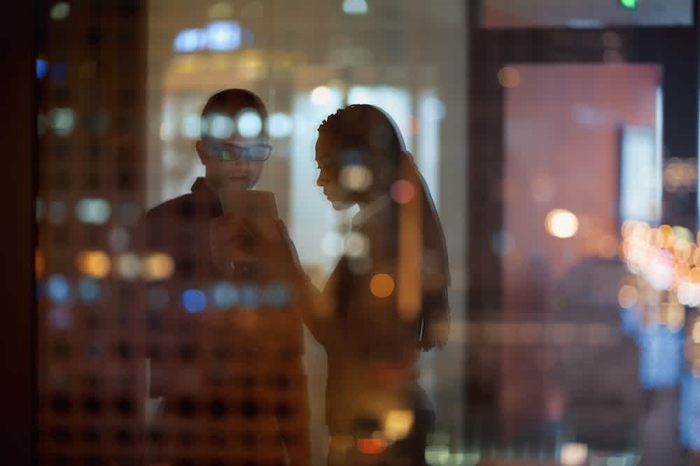 Two people stand indoors at night, looking at a tablet together. Their silhouettes are seen through a glass window, with blurred city lights and reflections creating a colorful, abstract effect.