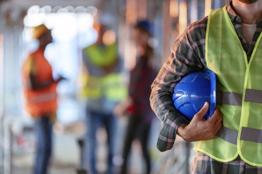 A close-up of a construction worker holding a blue hard hat, wearing a yellow safety vest, with three other workers in safety gear blurred in the background on a construction site.