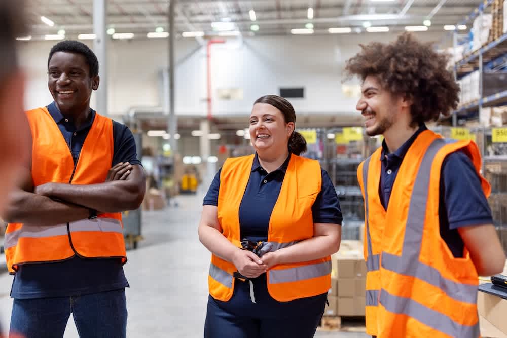 Three warehouse workers wearing orange safety vests stand together smiling and talking inside a large warehouse, with shelves and boxes visible in the background.