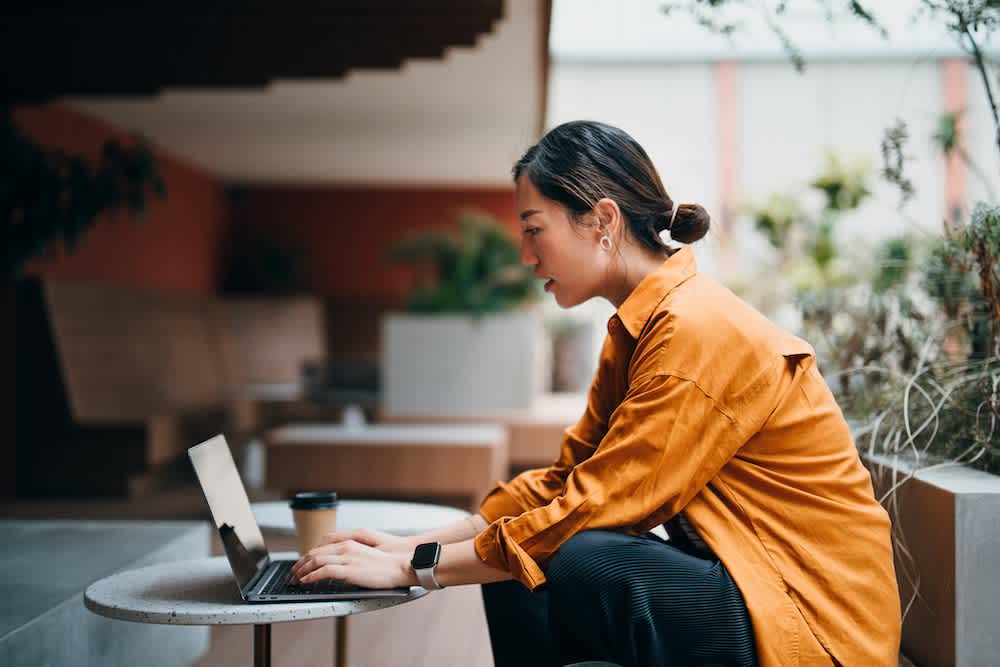 A woman in an orange shirt sits at an outdoor table, working on a laptop. A takeaway coffee cup is beside her, and she appears focused on the screen in a modern, open space with plants.