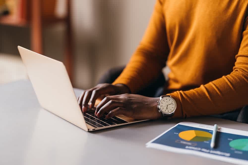 A person wearing an orange sweater types on a laptop at a desk, with a paper displaying colorful pie charts and a wristwatch visible on their left wrist.
