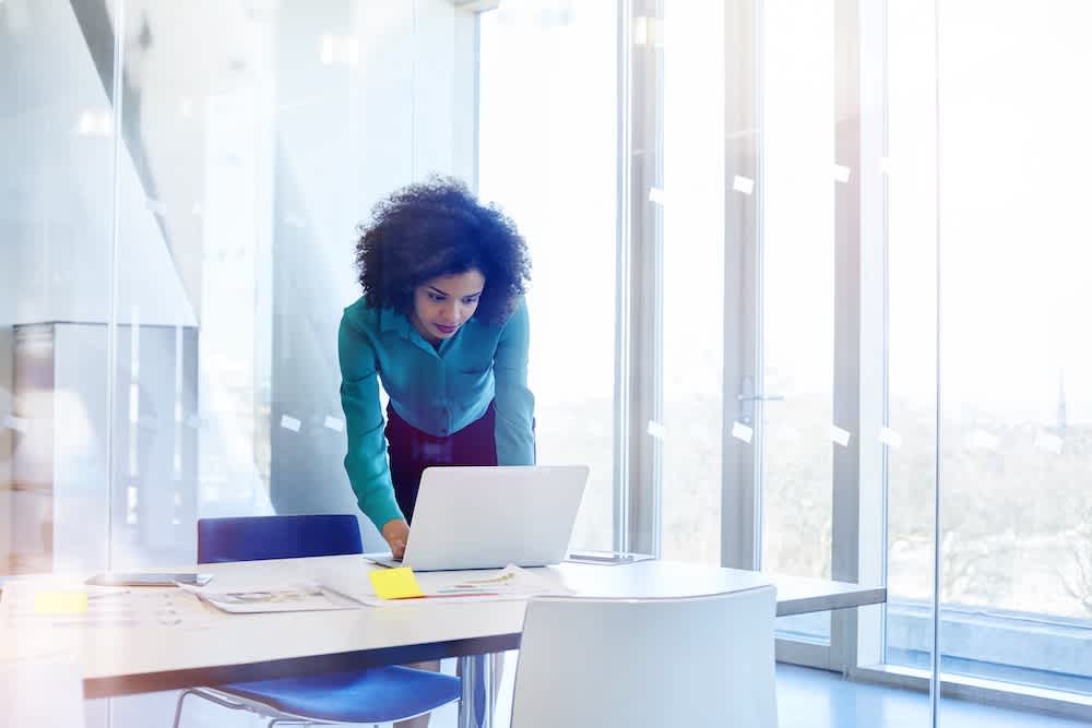 A woman with curly hair, wearing a teal blouse, stands and leans over a laptop on a desk in a bright modern office with large windows and scattered papers and sticky notes.