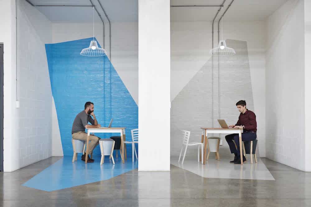Two men sit separately at desks in a modern office divided by a white wall. One side has a blue accent, the other is gray. Each man works on a laptop, creating a sense of isolation and symmetry.