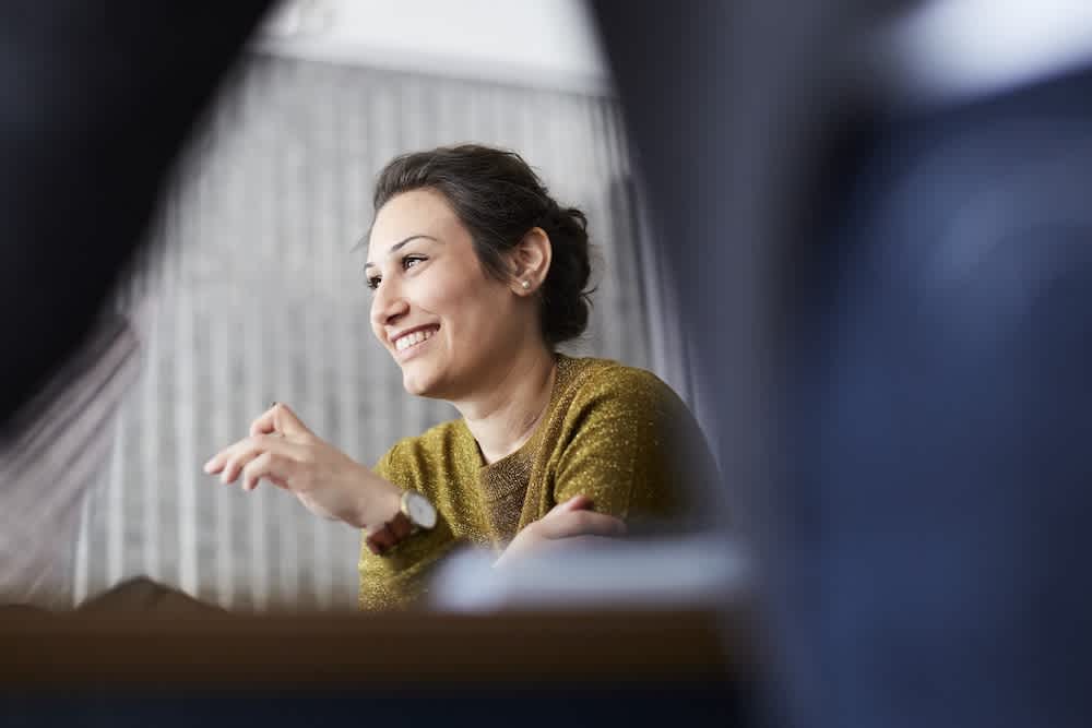 A woman with dark hair tied back, wearing a green sweater and a watch, sits at a table smiling and gesturing as she talks. The background is softly blurred.
