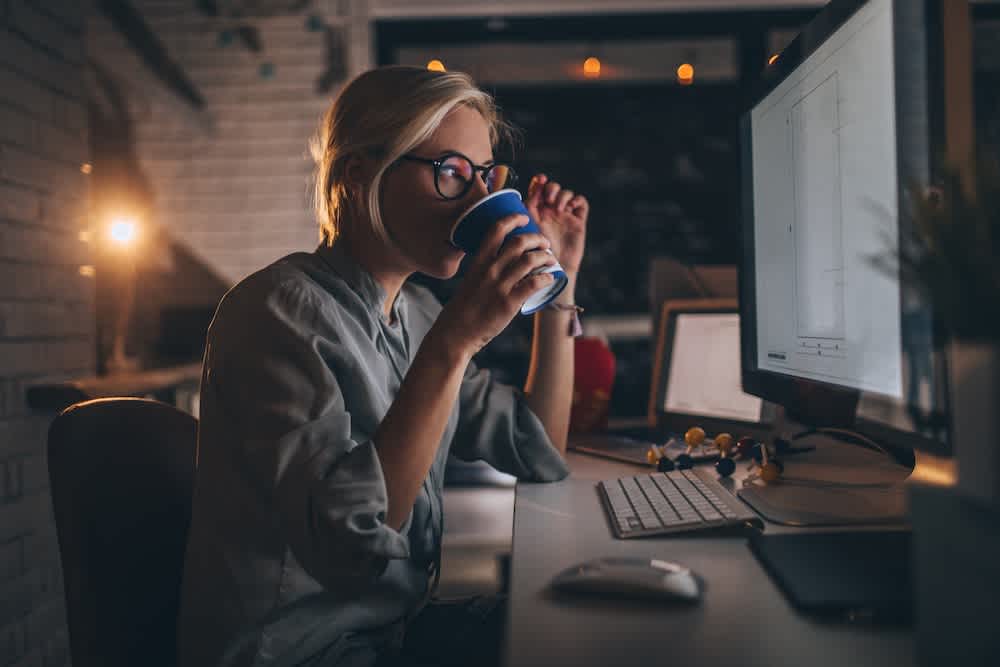 A woman wearing glasses sits at a desk in a dimly lit room, drinking from a blue mug while working on a computer. The workspace has a keyboard, mouse, and various small items.