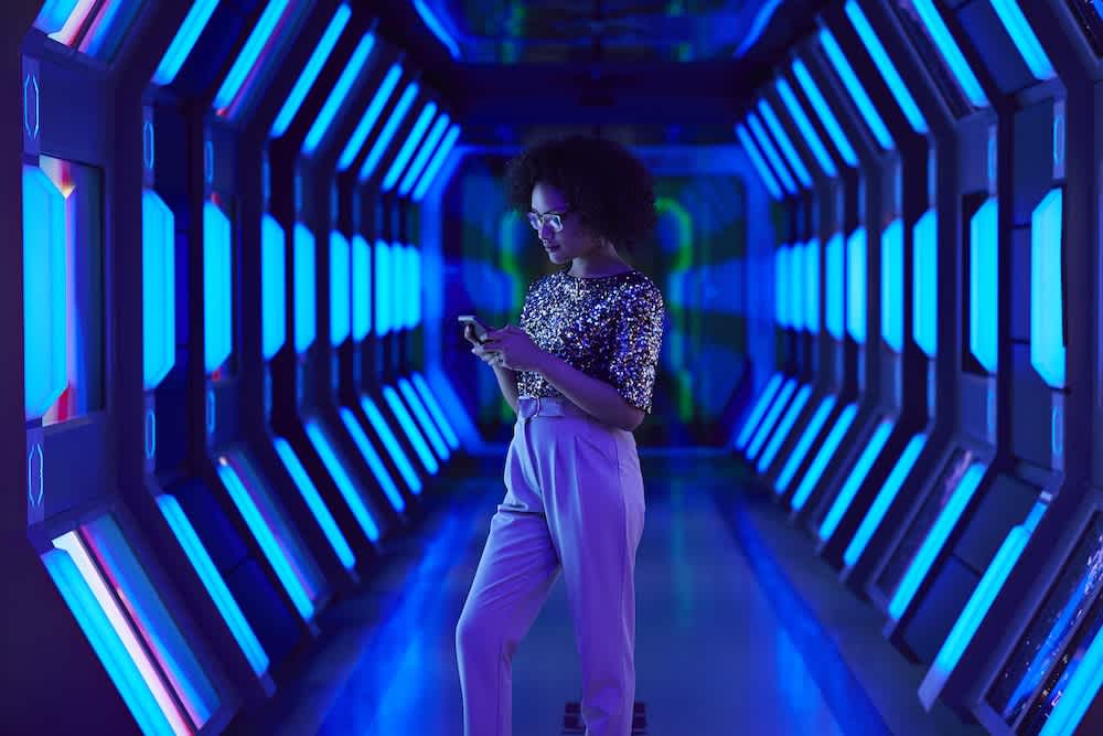A woman stands in a futuristic hallway with glowing blue and purple lights, looking at her phone. She wears glasses, a sequined top, and high-waisted pants. The corridor has a sci-fi, spaceship-like appearance.