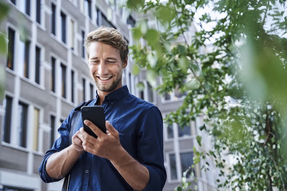 A smiling man in a blue shirt stands outdoors near a building and trees, looking at his smartphone.